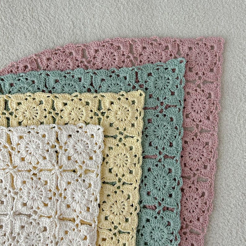 May include: A stack of four crocheted squares in varying pastel colors: white, pale yellow, mint green, and dusty rose. Each square features a floral pattern and a scalloped edge, showcasing a handmade craft.