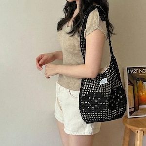 May include: A black crochet shoulder bag with an open-knit grid pattern and a small white rectangular label featuring black text. The bag has a wide strap and a soft, slouchy structure, worn over the shoulder to carry personal items.