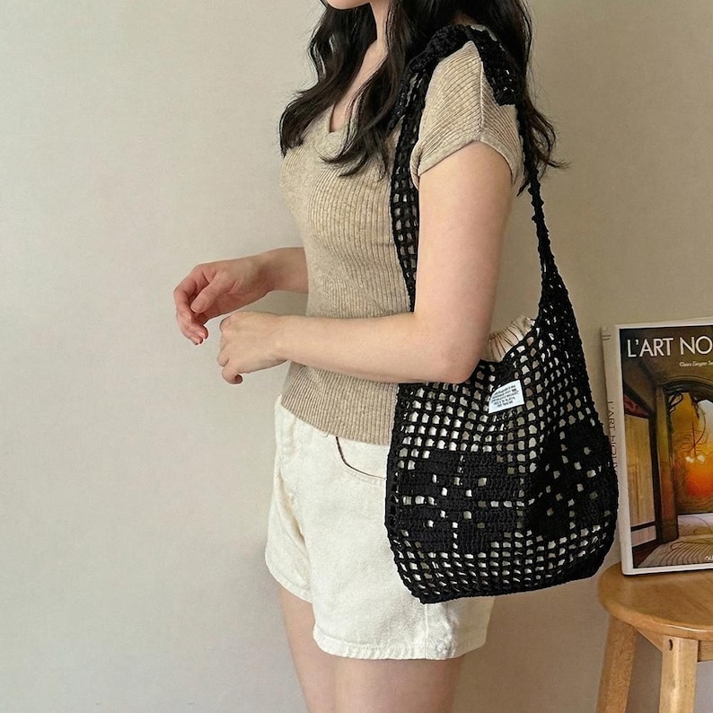 May include: A black crochet shoulder bag with an open-knit grid pattern and a small white rectangular label featuring black text. The bag has a wide strap and a soft, slouchy structure, worn over the shoulder to carry personal items.