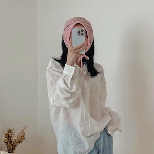 May include: A light pink crocheted bonnet with a delicate openwork design. The bonnet is tied under the chin with a matching pink string. The person is wearing a white button-down shirt and blue jeans.