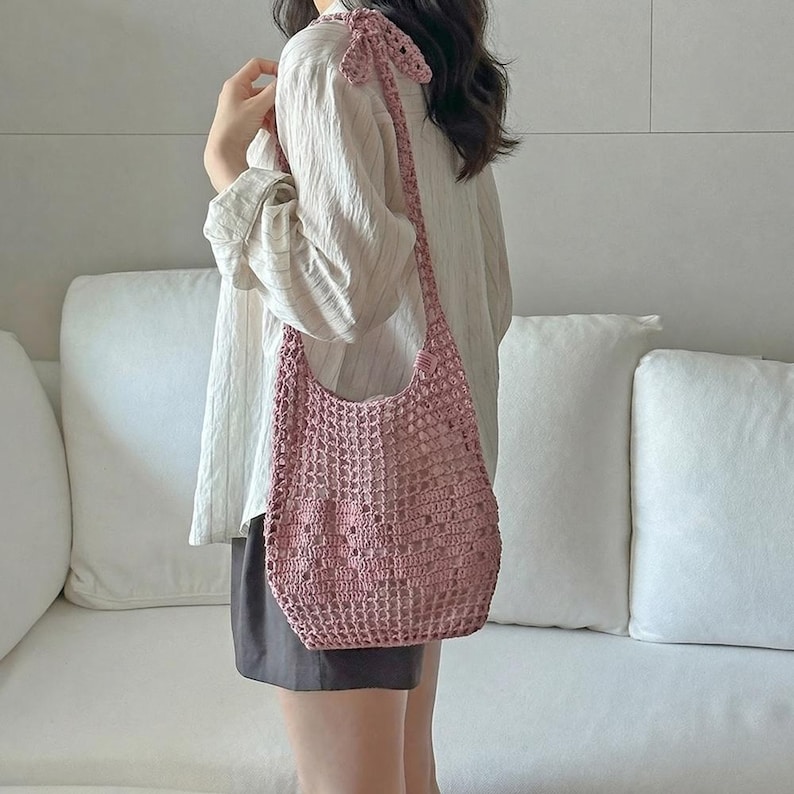 May include: A dusty rose crochet shoulder bag featuring an open-knit mesh pattern and a knotted strap detail. The bag is constructed from yarn with a visible grid-like texture, providing a flexible and lightweight structure for carrying items.