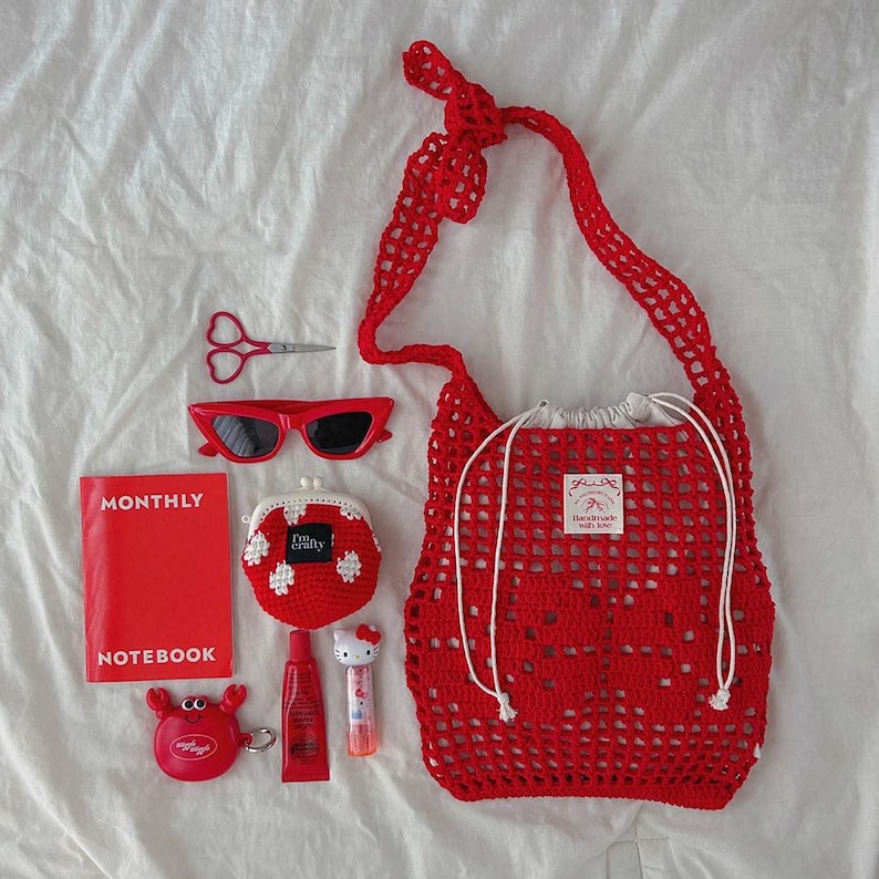 May include: A red crochet tote bag with a drawstring closure, a red monthly notebook, red sunglasses, red scissors, a red crochet coin purse, a red crab-shaped keychain, a red tube of lip balm, and a small white character-themed lip gloss.