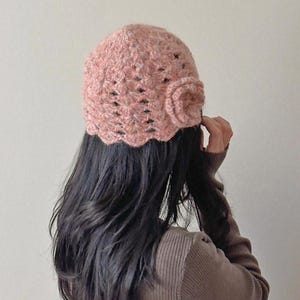 May include: A light pink crocheted hat with a scalloped brim and a flower detail on the side. The hat is made of a textured yarn and is worn by a person with long, dark hair. The hat is a stylish accessory.