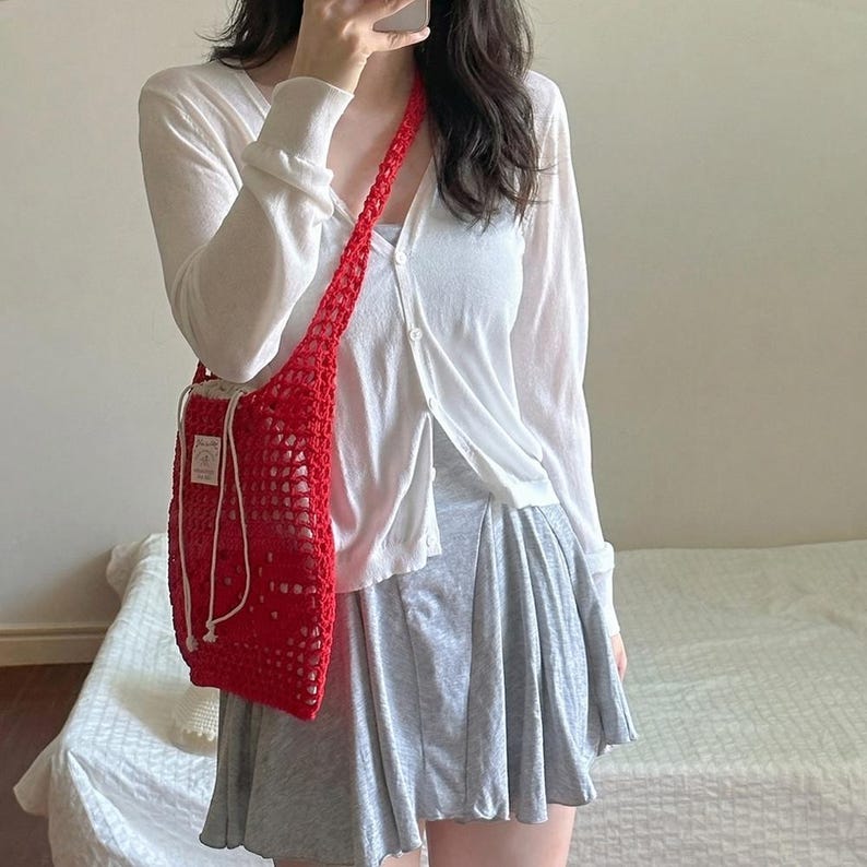 May include: A bright red crochet crossbody bag featuring an open mesh pattern, a long shoulder strap, and a white drawstring closure with a small rectangular label attached to the front. The bag is worn over a white long-sleeve button-down cardigan.