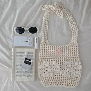 May include: A cream-colored crochet tote bag with a tie handle, white sunglasses, a Bobbi Brown card, two silver rings, a Sennok lip balm, a book with printed text, and a white digital music player with wired earbuds arranged on a white surface.