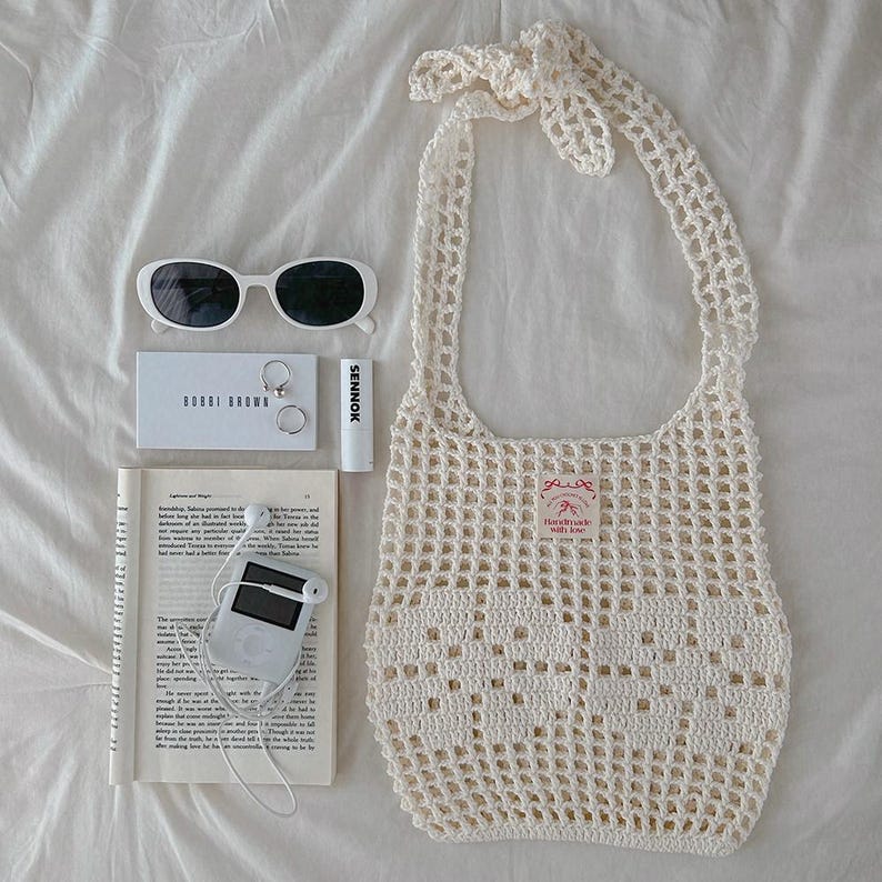 May include: A cream-colored crochet tote bag with a tie handle, white sunglasses, a Bobbi Brown card, two silver rings, a Sennok lip balm, a book with printed text, and a white digital music player with wired earbuds arranged on a white surface.
