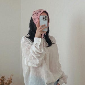 May include: A light pink, crocheted head covering with a delicate openwork design. The head covering is tied at the back and is worn with a white button-down shirt.