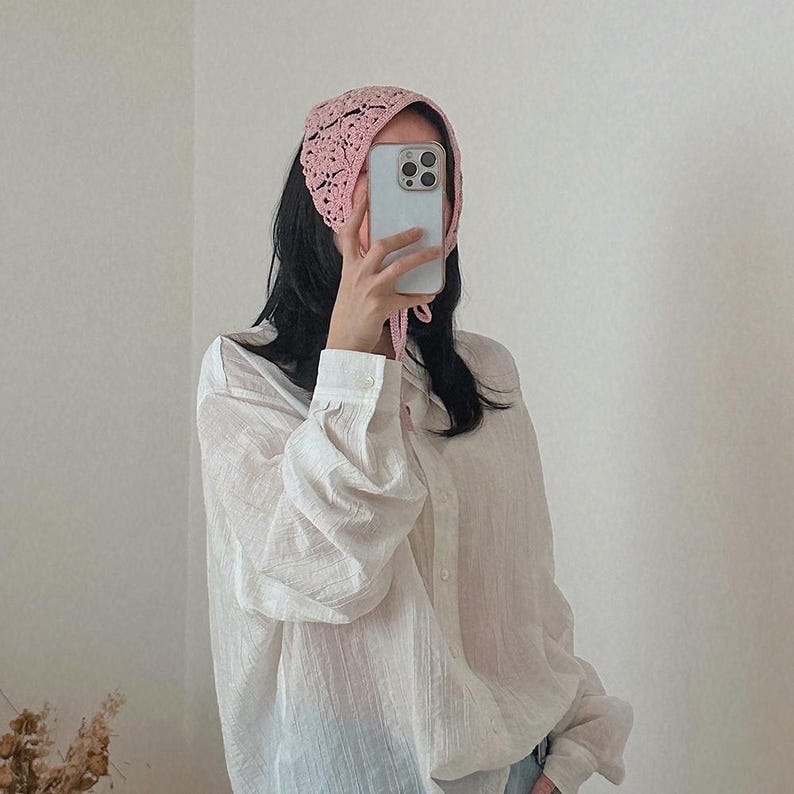 May include: A light pink, crocheted head covering with a delicate openwork design. The head covering is tied at the back and is worn with a white button-down shirt.
