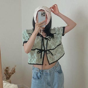 May include: A light green, short-sleeved top with black trim and a bow closure, paired with light blue jeans. A white, lace bonnet is being adjusted. The top has a textured floral pattern.