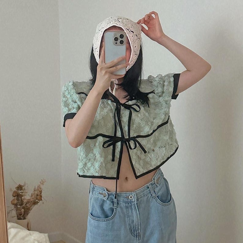 May include: A light green, short-sleeved top with black trim and a bow closure, paired with light blue jeans. A white, lace bonnet is being adjusted. The top has a textured floral pattern.