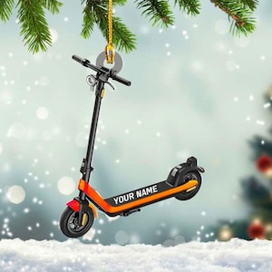 May include: A black, orange, and red scooter ornament with the text "YOUR NAME" on the side. The scooter is hanging from a gold string with a silver disc, with a blurred background of a Christmas tree and snow.