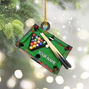 May include: A miniature pool table ornament with a green felt surface, a rack of billiard balls, and two pool cues. The ornament has the text "YOUR NAME" on the felt. It is suspended by a gold string.