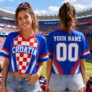 A smiling woman wearing a blue and red Croatia soccer jersey with the number 00, standing in front of a soccer stadium.