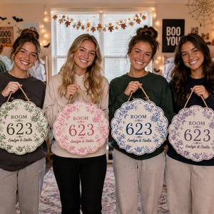 May include: Four round, scalloped-edge wooden signs with floral borders, each featuring the text "ROOM 623" and "Evelyn & Stella" in the center. The signs are displayed in green, pink, blue, and purple color variations, each with a jute rope hanger.