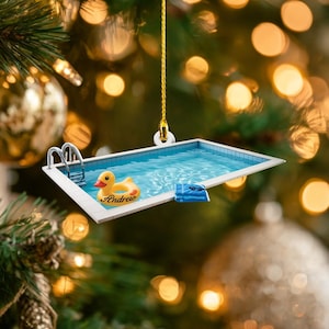 May include: A Christmas tree ornament shaped like a swimming pool. The pool has blue water, a yellow rubber duck with the name "Andrew", and a blue towel. The ornament is hanging from a gold string.