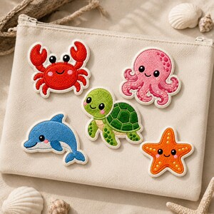 May include: A beige canvas zippered pouch featuring five embroidered sea creature patches: a red crab, a pink octopus, a green sea turtle, a blue dolphin, and an orange starfish. Each patch has a smiling face and detailed stitching.