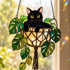 May include: A stained glass suncatcher featuring a black cat sitting inside a beige macrame-style hanging planter. The planter is surrounded by green monstera leaves and is suspended by two black metal chains, creating a decorative window piece.