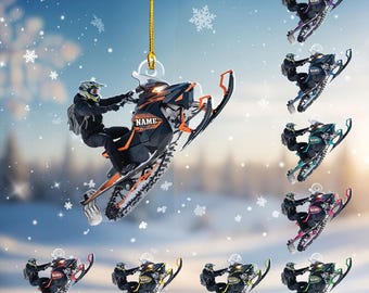May include: A clear acrylic ornament featuring a snowmobile with a rider, accented with orange and black details. The word "NAME" is visible on the snowmobile. Additional ornaments in various colors are also shown.
