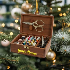 May include: A wooden sewing kit ornament with a hinged lid, containing spools of colorful thread, scissors, and other sewing tools. The box is engraved with the text "Your Name" in yellow. The ornament hangs from a gold string against a Christmas tree background.