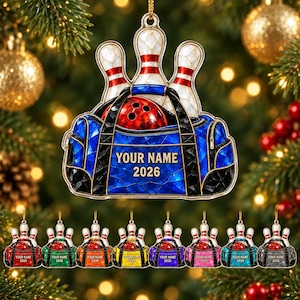 May include: A decorative hanging ornament featuring a blue bowling bag with a red bowling ball and three white bowling pins with red stripes. The bag displays the text YOUR NAME and 2026. The design has a stained glass effect with gold trim.