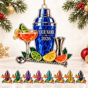 May include: A decorative hanging ornament featuring a blue cocktail shaker with the text YOUR NAME and 2026, a cocktail glass with orange liquid, a jigger, a mixing spoon, and slices of lime, lemon, and orange. A row of smaller versions is below.