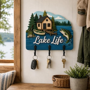 May include: A wooden wall-mounted key holder featuring a layered design of a cabin, a boat, and a fish on a blue lake background. The text "Lake Life" is printed in white script. Three black metal hooks hold various metal keys and a car key fob.