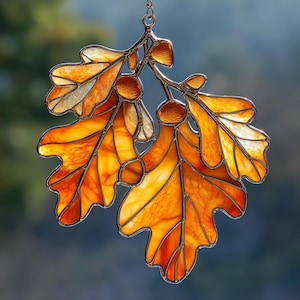 May include: A stained glass suncatcher featuring two oak leaves and two acorns in shades of amber, orange, and yellow. The glass pieces are held together by thin metal soldering, forming a detailed botanical design with a hanging loop at the top.