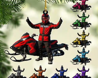 May include: A Christmas ornament featuring a snowmobile and a person in a red and black snowmobile suit with the name "Henry". The ornament is hanging from a gold string. Additional ornaments of different colors are also shown.