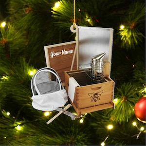 May include: A Christmas tree ornament featuring miniature beekeeping equipment. The ornament includes a wooden beehive with a bee graphic, a smoker, a beekeeper's veil, and a wooden frame with the text "Your Name".