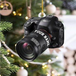 May include: A black camera ornament with the text "YOUR NAME" and "2025" on the lens. The ornament is hanging from a gold string, with a blurred Christmas tree background.