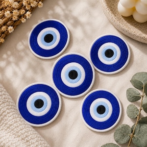 May include: Five round, embroidered patches featuring a blue evil eye design. Each patch has a dark blue outer ring, a white inner ring, a light blue ring, and a black center. The patches are arranged on a neutral-colored surface.