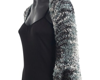Könnte beinhalten: Ein durchsichtiges Bolero mit offenem Gewebe und Dreiviertelärmeln. Das Bolero ist eine Mischung aus Silber, Grau und Schwarz, wodurch ein strukturiertes, schimmerndes Aussehen entsteht. Das Kleidungsstück wird auf einer schwarzen Schaufensterpuppe präsentiert.
