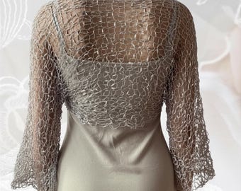 Könnte beinhalten: Ein silberner, metallisch glänzender Bolero mit einem offenen, grob gestrickten Häkelmuster und langen Ärmeln. Das Kleidungsstück wird über einem einfarbigen, hellen ärmellosen Kleid getragen. Der Bolero hat eine netzartige Textur mit Schimmer.