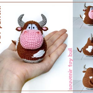 May include: A handmade crochet bull toy with a brown body, white patches, and a pink snout. The bull has gray horns and is held in a hand. The image also includes the text "PDF pattern" and "souvenir toy bull".