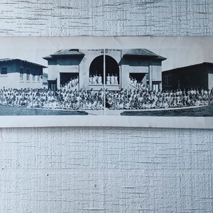 May include: A black and white panoramic photograph of a school group posing in front of a building. The image shows a large group of people, likely students, seated on the grass. The school building has a central arched entrance and is flanked by other structures.