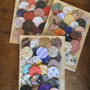 Könnte beinhalten: Drei Packungen Stoff-Yo-Yos in verschiedenen Farben und Mustern, darunter Blumen-, Streifen- und unifarbene Stoffe. Die Yo-Yos sind auf einem Vintage-Papierhintergrund angeordnet und in klarem Kunststoff versiegelt.