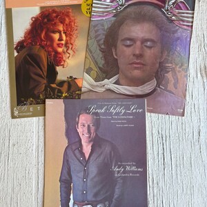 Può includere: Tre copertine di spartiti musicali con un uomo e una donna sulla copertina. Le copertine sono per le canzoni "From a Distance", "Dreamweaver" e "Speak Softly Love (Love Theme from The Godfather)".