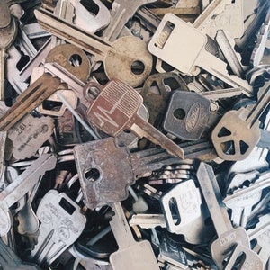 May include: A collection of assorted metal keys, viewed from above. The keys vary in size and design, with a mix of silver and grey tones, and some showing signs of age. Some keys have text and markings. The image presents a textured, detailed composition.