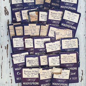 Puede incluir: Una colección de etiquetas vintage de piezas de reloj, principalmente moradas con texto blanco, para relojes Elgin, Hamilton, Waltham e Illinois. Las etiquetas son para muelles, y algunas incluyen el número de fábrica y el tamaño. Las etiquetas están dispuestas en un patrón aleatorio sobre una superficie de madera blanca.