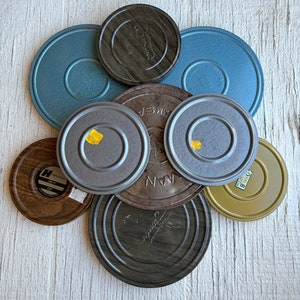 May include: A collection of eight vintage film reels in various colors and materials, including blue, brown, silver, and gold. The reels are arranged in a circular pattern on a white wooden surface.
