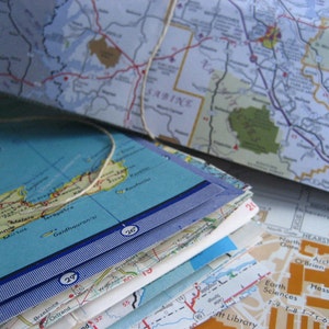 Può includere: Un mazzo di mappe piegate con colori blu, bianco e verde. Le mappe sono etichettate con nomi di luoghi e numeri. La mappa superiore è piegata sulle altre e mostra una mappa di una zona costiera con un mare blu e una massa terrestre verde. Le mappe sono legate insieme da uno spago bianco.