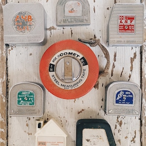 Op de afbeelding: Een assortiment vintage meetlinten in verschillende kleuren en maten, waaronder een rode meetlint met de woorden "Roe Comet" en "Steel Measuring Tape" erop gedrukt, een witte meetlint met de woorden "Luerin White Clad" erop gedrukt en een zilveren meetlint met de woorden "K&E Wyteface LST Level Square Tape" erop gedruckt.