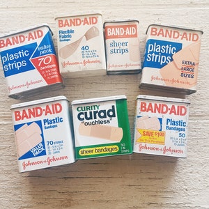May include: A collection of vintage Band-Aid tins, featuring various sizes and types of bandages, including plastic strips, sheer strips, and flexible fabric bandages. The tins are all white with red and blue lettering and feature the Johnson & Johnson logo.