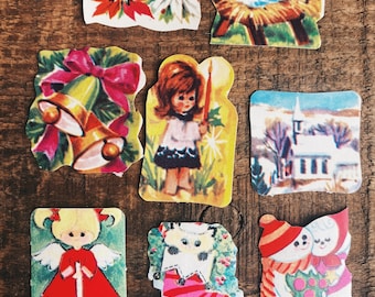 Könnte beinhalten: Ein Set mit Vintage-Weihnachtsaufklebern mit verschiedenen weihnachtlichen Motiven, darunter eine Weihnachtsstern, eine Krippe, eine Kirche, ein Schneemann, eine Katze und Engel. Die Aufkleber sind auf weißem Papier mit bunten Illustrationen gedruckt.