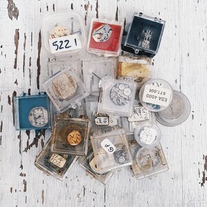 May include: A collection of small, clear plastic boxes containing various watch parts, including gears, hands, and a small watch face. The boxes are labeled with numbers and text, including "ESA 4000", "977.001", and "MURSE DENTAL LABORATORY".