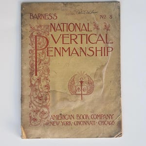 May include: A vintage book cover with a tan background and red text. The title is "National Vertical Penmanship" with the author's name "Barnes's" and the publisher "American Book Company" printed below. The book cover features a decorative border with a stylized image of a torch and wings.