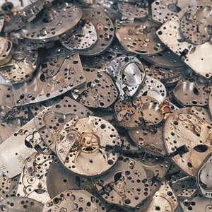 Puede incluir: Primer plano de una colección de piezas de relojes antiguos. La imagen muestra una variedad de piezas metálicas plateadas y grises, incluyendo engranajes, placas y otros mecanismos. Algunos componentes tienen detalles dorados. Las piezas son de varias formas y tamaños.