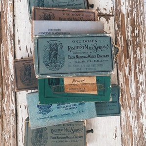 May include: A stack of vintage boxes containing watch parts. The boxes are green, brown, and blue with white lettering. The text on the boxes reads "One Dozen Resilient Main Springs Manufactured by Elgin National Watch Company Elgin, Ill, U.S.A."