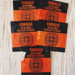 Puede incluir: Cinco paquetes naranja y negro de cuerdas de guitarra Yamaha Dynamic. Cada paquete tiene un tamaño de cuerda diferente etiquetado como A5, E6, D4, G3 y B2.