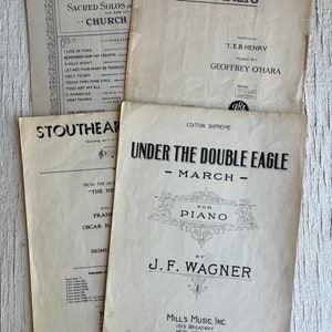 Puede incluir: Una portada de partitura de música vintage para una marcha de piano titulada "Bajo el doble águila" de J.F. Wagner. La portada está impresa en tinta negra sobre papel blanco y presenta un borde decorativo.